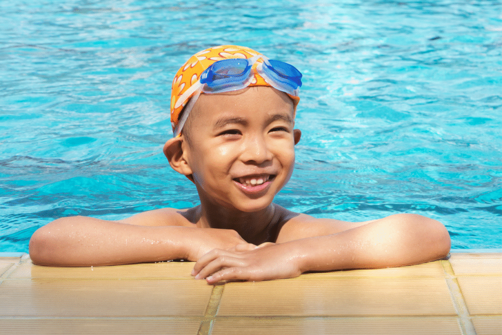 Child in googles and swim caps with arms up on the side of swimming pool.